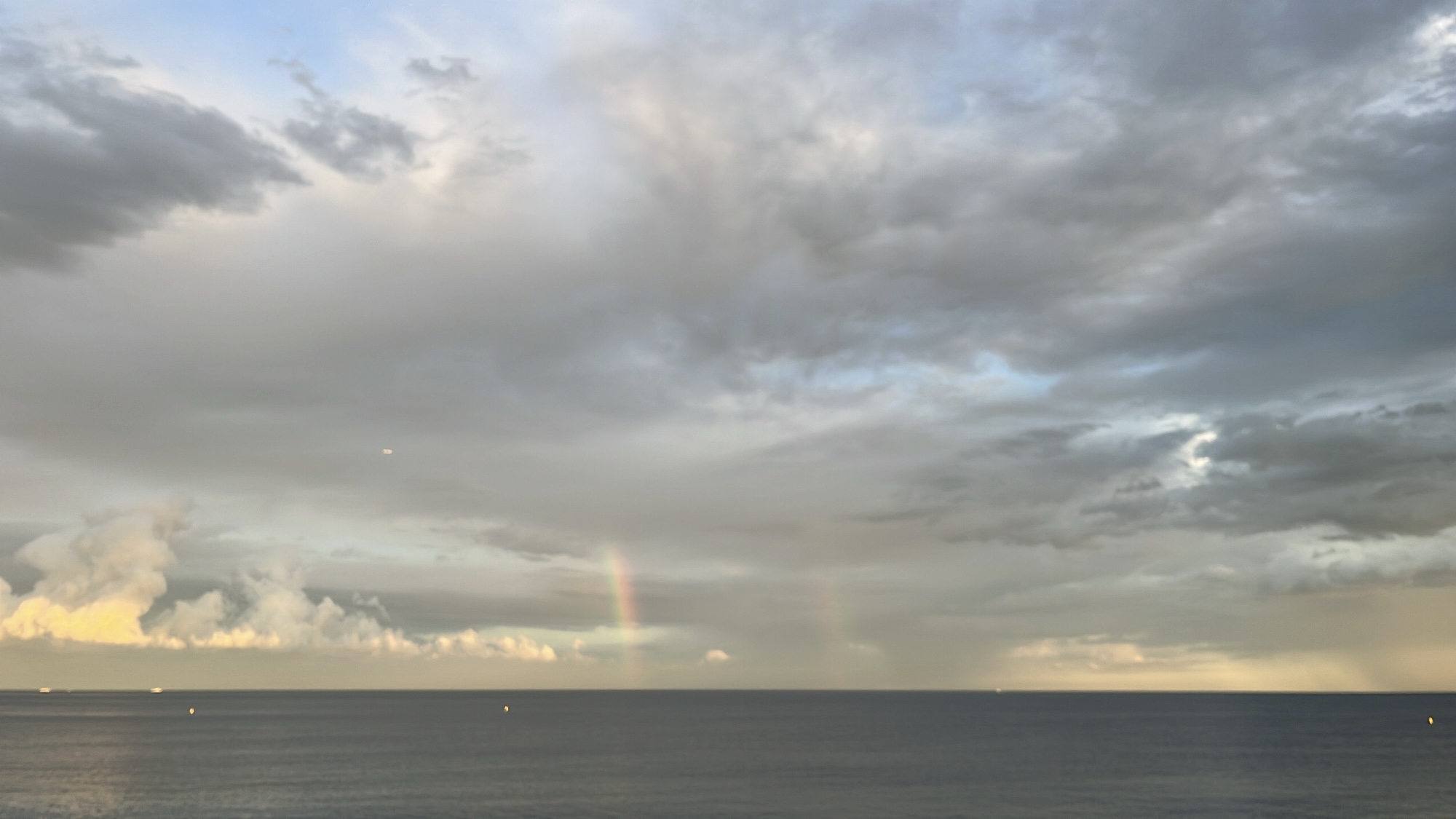 Faint rainbow over a still sea under soft cloud cover at dusk